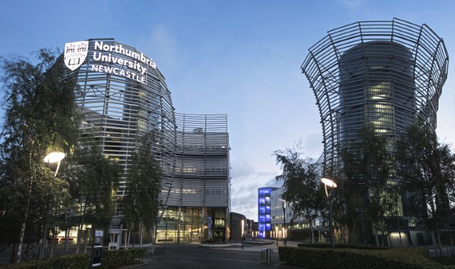 This is a photograph of two buildings that are curved in shape with lots of grey and glass visible. There is a pathway between the two buildings and trees on each side of the pathway. On one of the buildings are the words 'Northumbria University NEWCASTLE' and a shield logo, both in white.