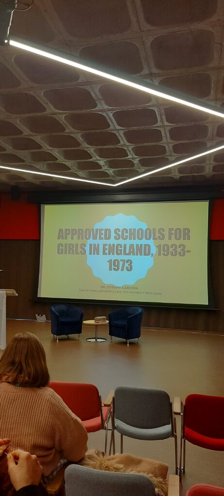 The image is a photograph that shows the inside of a lecture theatre. The focal point is a big screen that has a presentation visible. It says APPROVED SCHOOLS FOR GIRLS IN ENGLAND, 1933-1973 in a dark bold font, on a primarily yellow background with a white shape in the centre. In front of the screen are two chairs and a table with books on. The view is taken from the audience, and it is possible to see some of the chairs and attendees looking on.