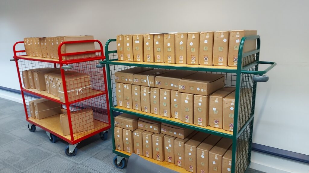 This image is a photograph of the inside of the National Archives reading room, and there are two trollies loaded with J 77 box files. One trolley is green, and the other is red. Both have three shelves that have multiple brown box files on them.