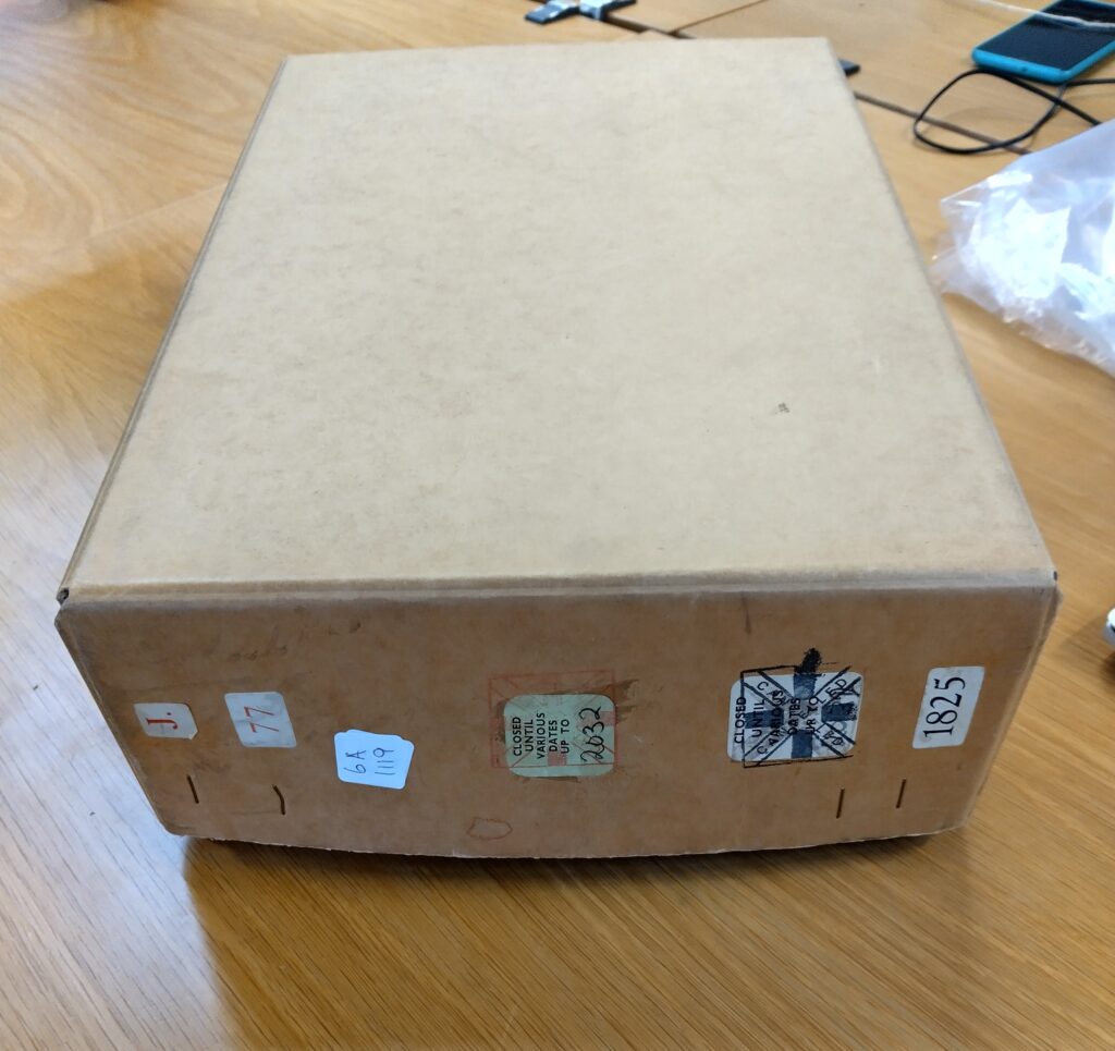 The image is a photograph of a rectangular box file made of thick brown cardboard, sat on a desktop made of light wood. On the front of the box are multiple labels including one saying J 77 another with a cross stamped on it that says ‘CANCELLED’ underneath this is a date when the files would be opened for public view. There is also a label with the box number, in this case 1825.